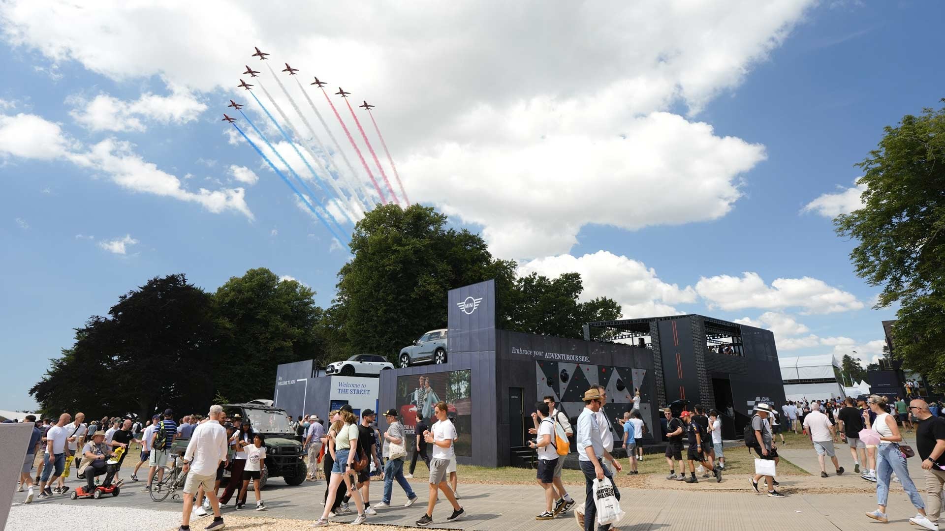 Airshow with jets emitting red, white, and blue trails in a clear sky. Below, a bustling outdoor event with people walking near a modern exhibit. Atmosphere: festive.